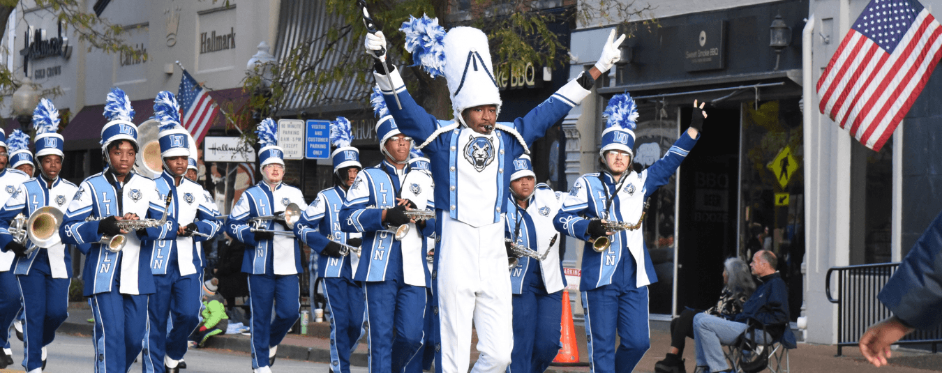 A drum major leads Lincoln University of Missouri’s marching band during a downtown parade, raising both arms as band members perform behind them.