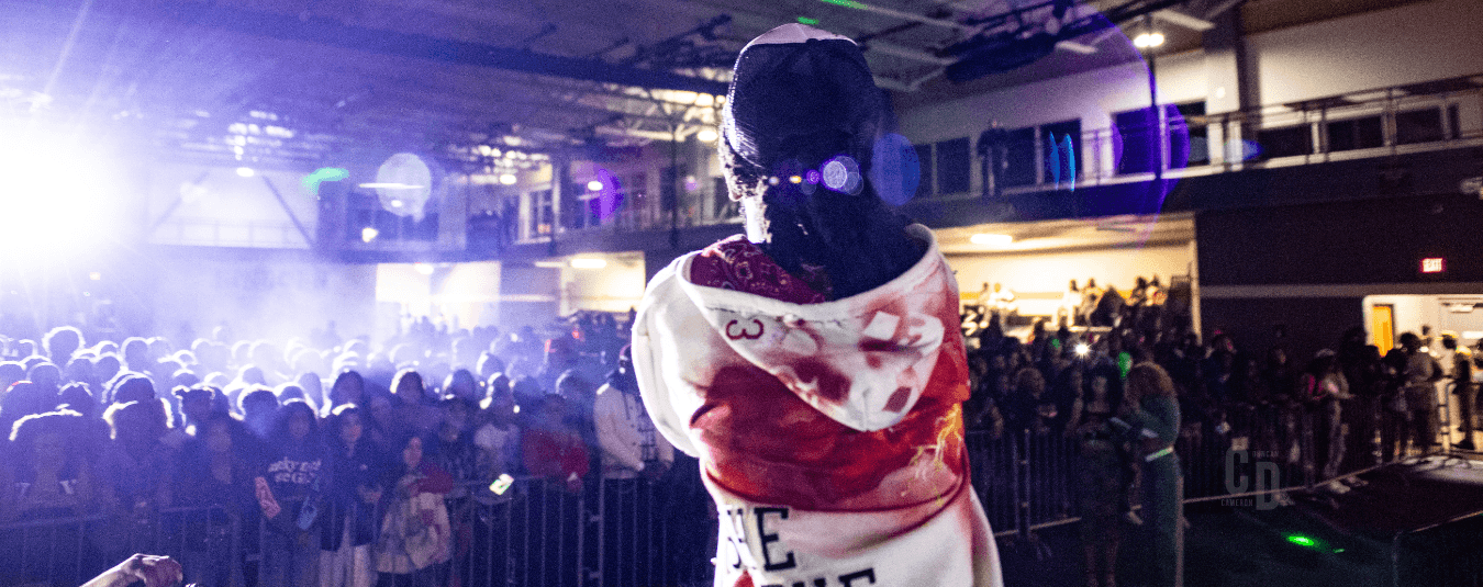A performer stands on stage facing a large crowd during a high-energy indoor concert attended by students at Lincoln University of Missouri.
