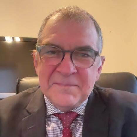 Headshot image of Dr. Adrian Petrescu. He wears a glasses, a suit and tie, and is seen against a yellow/beige background. 