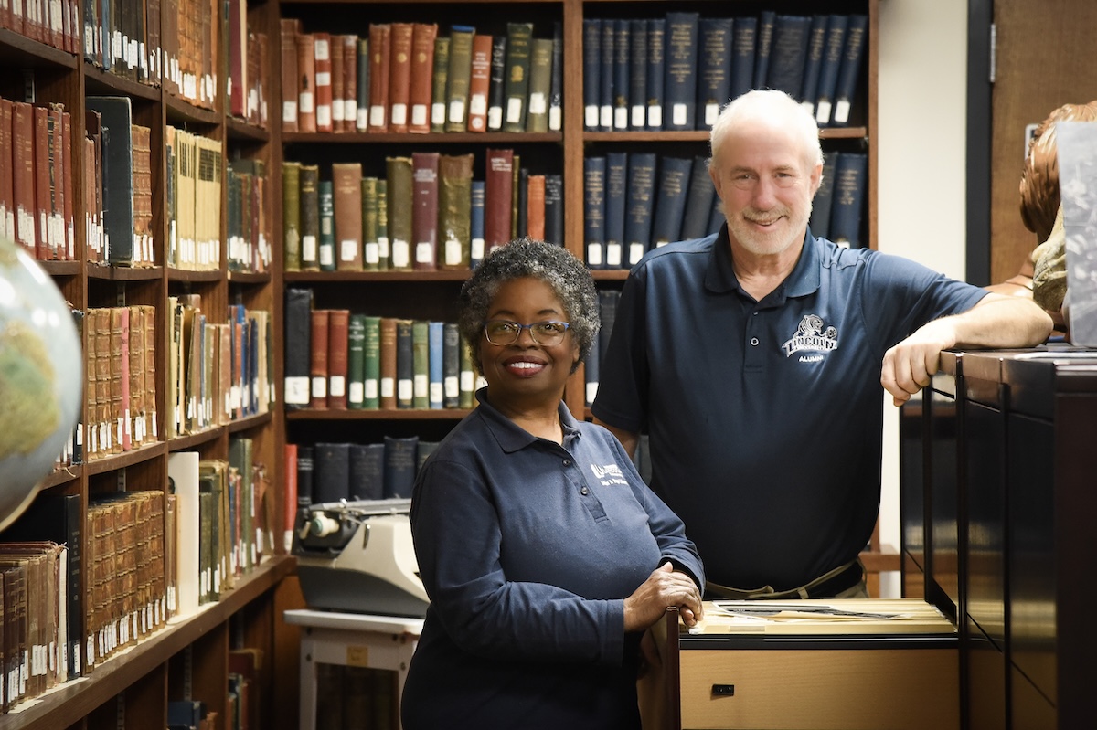 A smiling woman and man, both wearing navy blue polo shirts with a Lincoln University logo, stand in an archive room filled with bookshelves. The woman is in the foreground, leaning on an open file cabinet drawer, while the man stands slightly behind her, resting his arm on the cabinet. The background features shelves lined with vintage books, a typewriter, and a globe.
