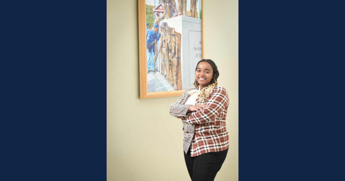 Smiling woman standing with arms crossed in front of a framed artwork on a light-colored wall." Let me know if you'd like to include details like her clothing style or the setting (e.g., a campus or office).