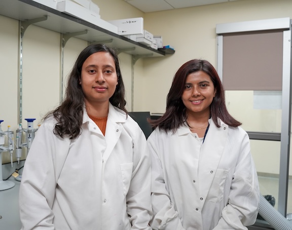 Two young women in lab coats stand in a small laboratory. 