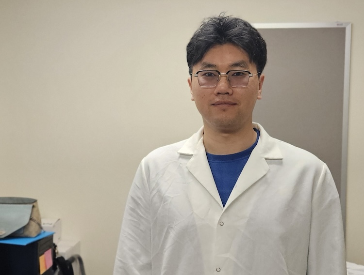 : A young man in glasses and a lab coat stands in a small laboratory. 
