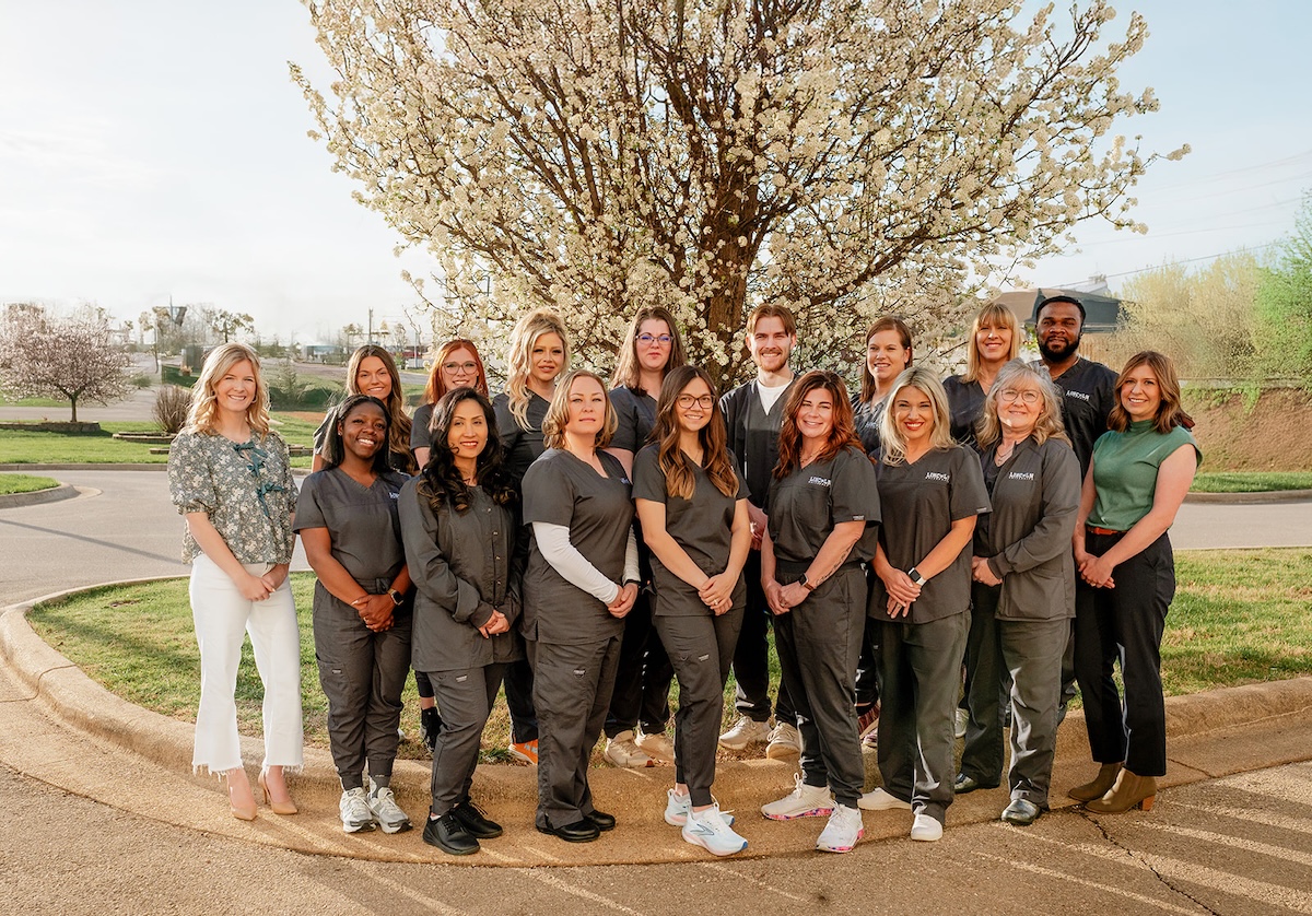 A group of nursing graduates and faculty from Lincoln University of Missouri pose together outdoors in front of a blooming tree during spring. Most are wearing gray scrubs, while a few are dressed in business casual attire. They are smiling and standing in two rows on a circular curb under a clear sky.