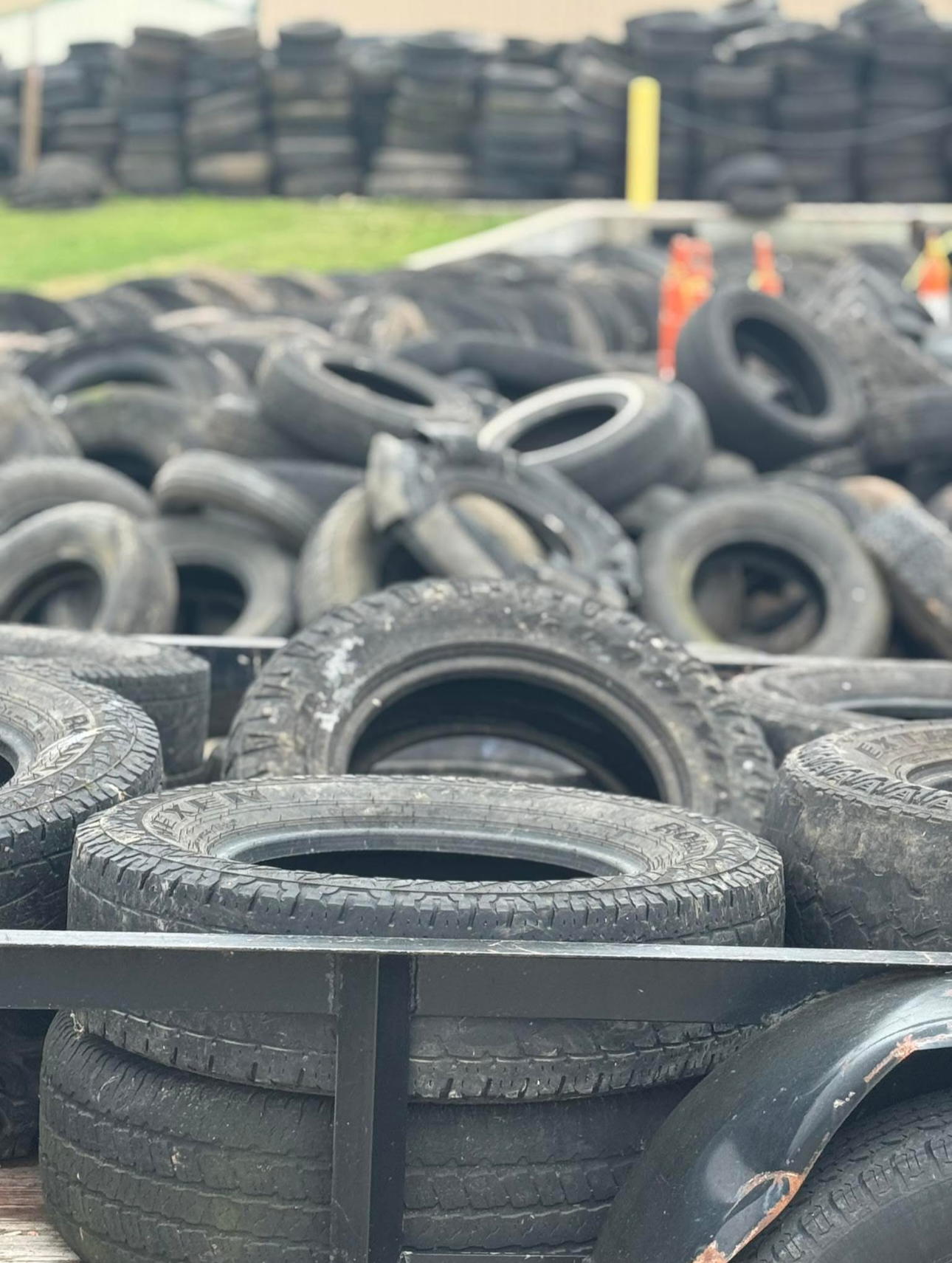 A pull-behind trailer is full of tires and sits behind piles of more tires.