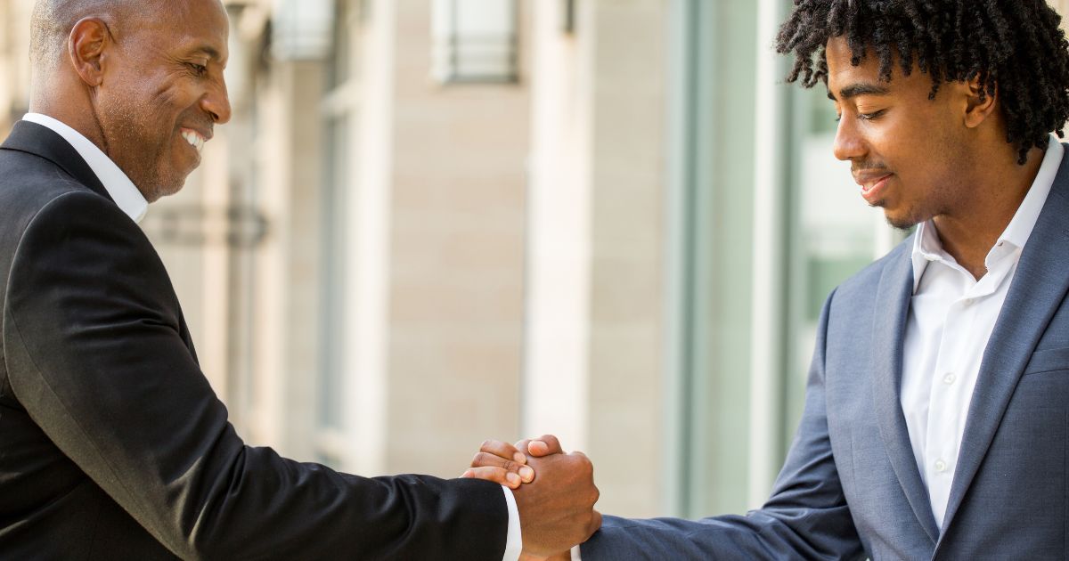 Two men in business attire share a handshake outside a building, symbolizing mentorship, networking, or professional connection.