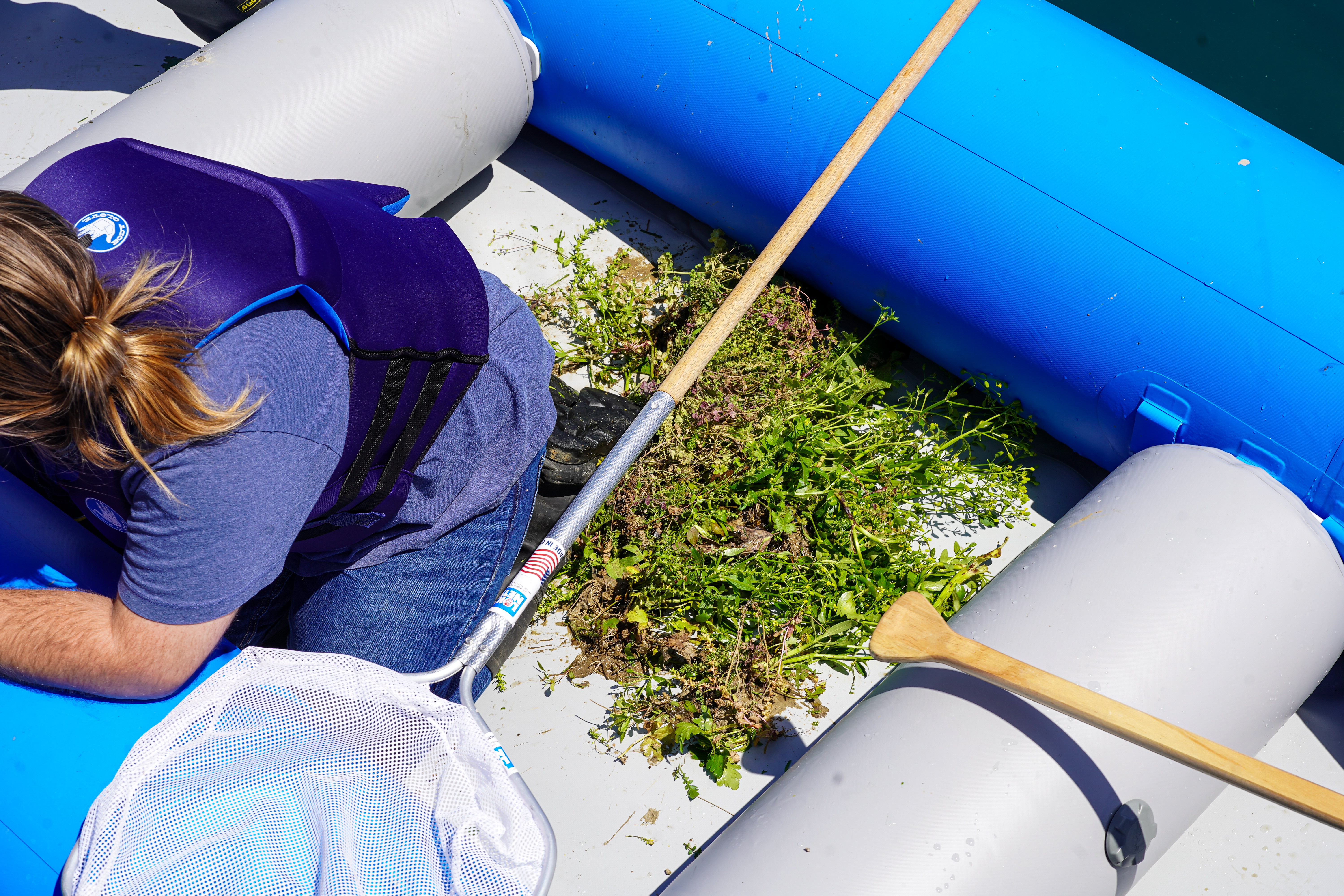 A young man crouches in an inflatable boat filled with picked weeds and grass.