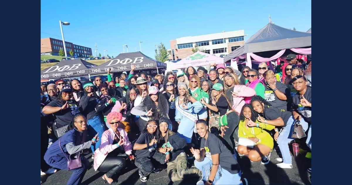 A large group of women pose together outdoors at a lively event, smiling and making hand signs associated with their sorority. Many wear coordinated outfits in green, pink, black, and denim, with branded shirts, jackets, and accessories. Behind them are tents labeled with Greek letters “ΔΦΒ” and decorated with pink drapes. The setting appears to be a college campus with academic buildings and a bright blue sky in the background, suggesting a celebration or homecoming event.