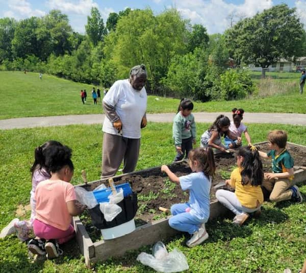 Children gather around a raised garden bed, planting seeds with guidance from an Urban Green Dreams. 