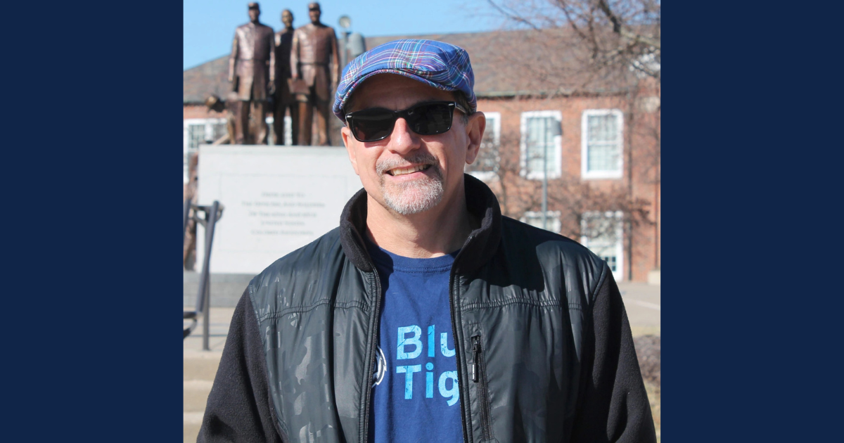 A man wearing sunglasses, a plaid flat cap, and a black jacket stands smiling in front of a monument featuring four bronze statues. He is outdoors on a sunny day, with a red-brick building and bare trees in the background.