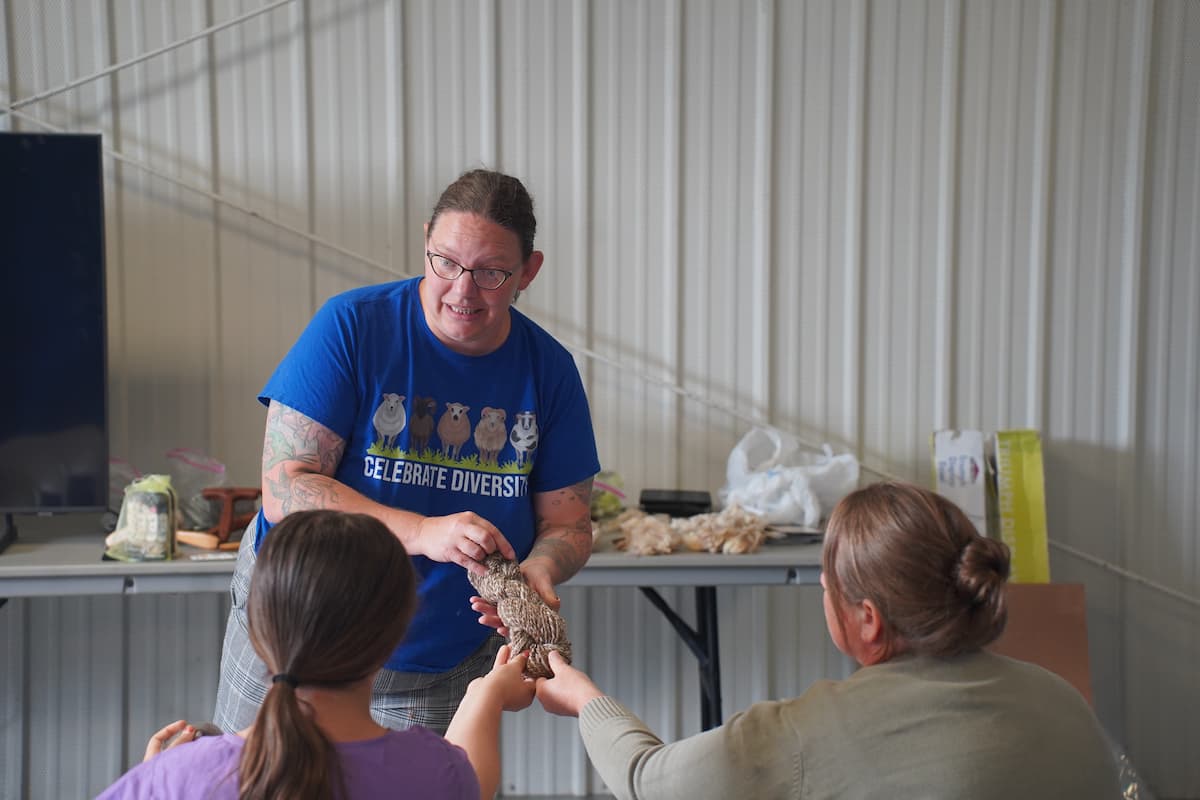 A woman holds out a bundle of spun wool for two sitting audience members to touch.