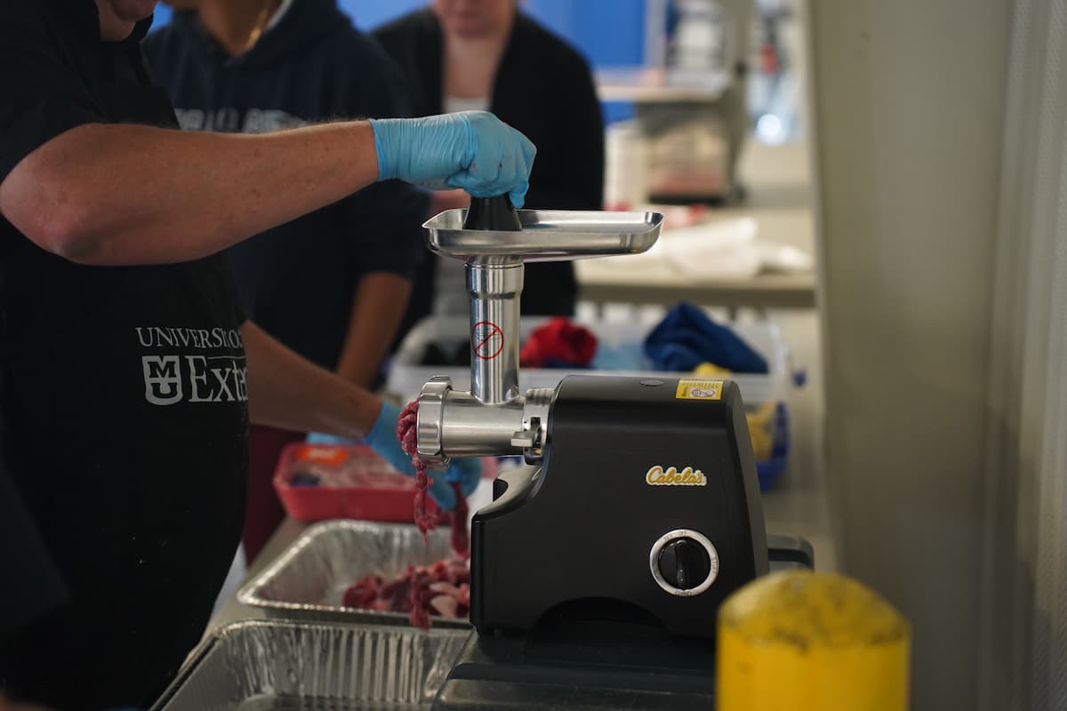 A man pushes goat meat into a grinder, which pushes ground meat into a tray.