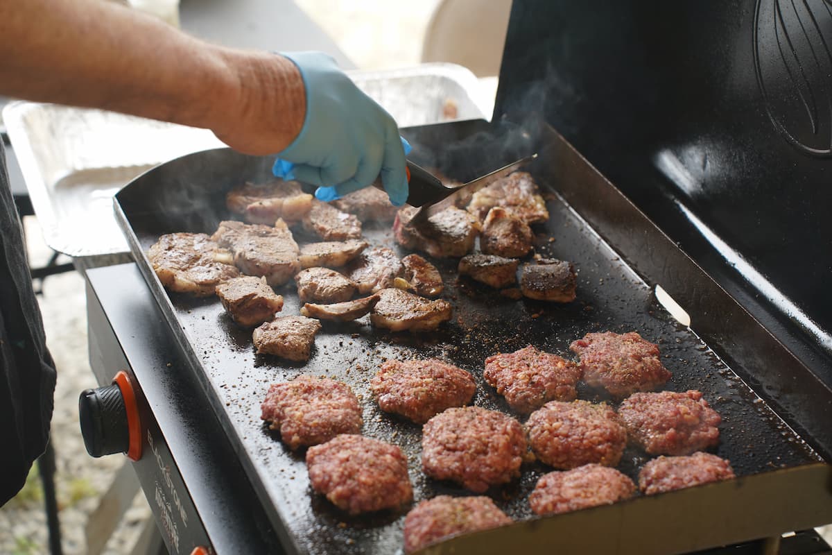 A man cooks several goat burgers on an outdoor grill.