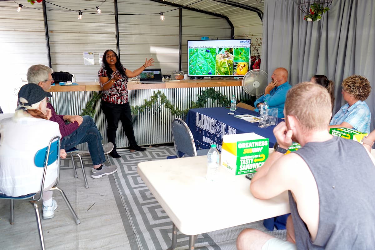 A woman stands in a room and gives a presentation off of a screen to a small audience.