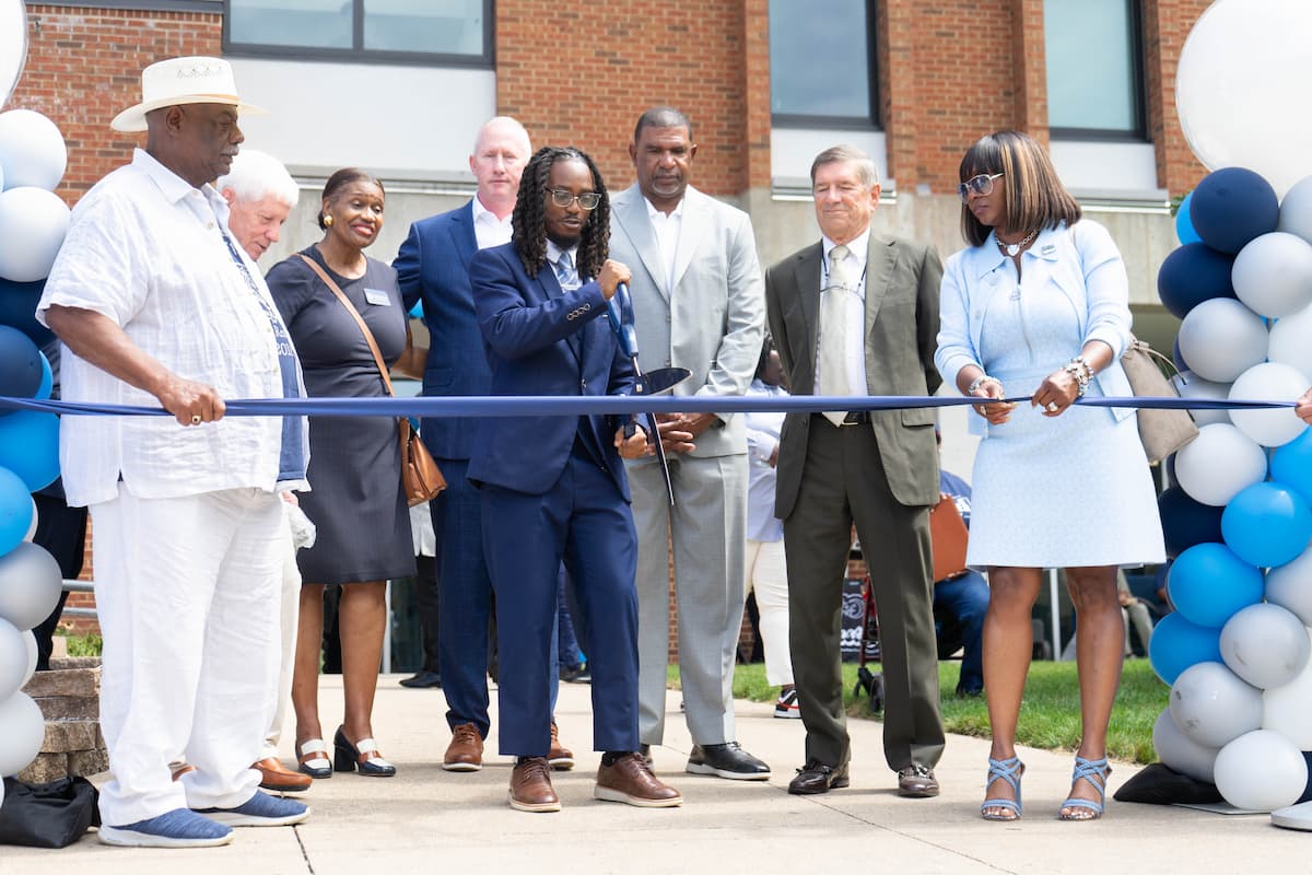Group cuts blue ribbon at ceremony in front of brick building, with balloons decorating the entrance.