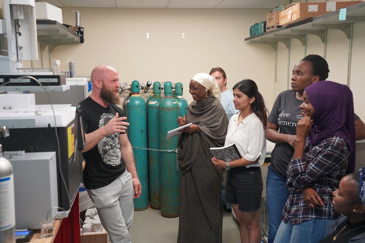 A man speaks to a group of six young adults in a small laboratory. 