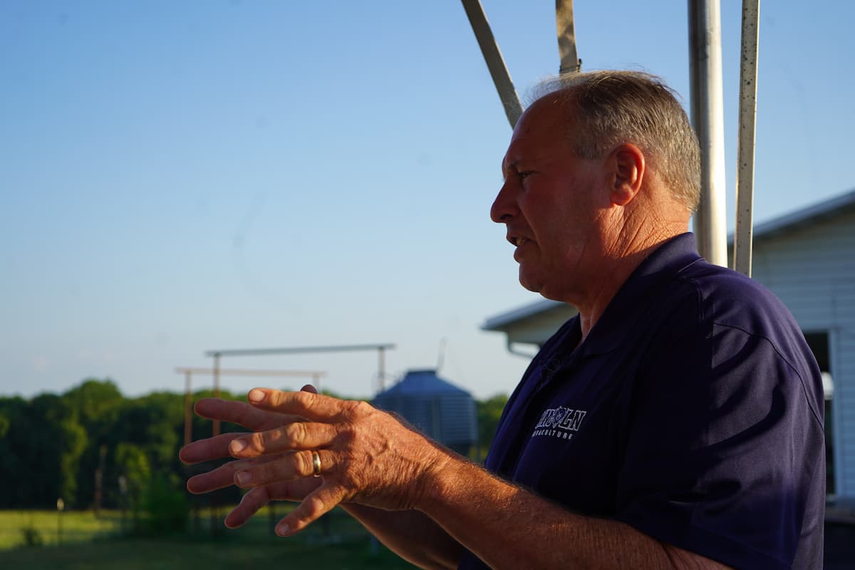 A man in a dark blue shirt speaks to an unseen audience on a tractor-pulled trolley. 