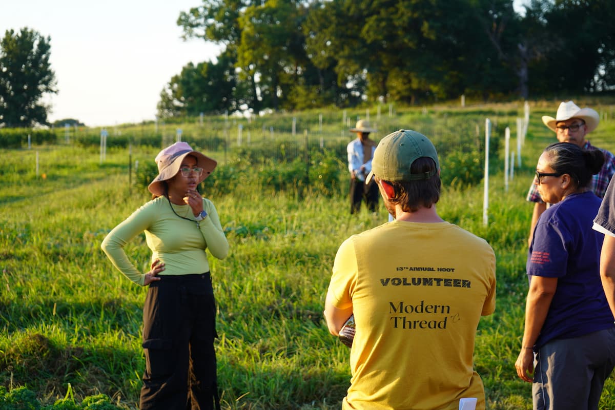 A young woman stands in a row of crops speaking to several people in front of her.