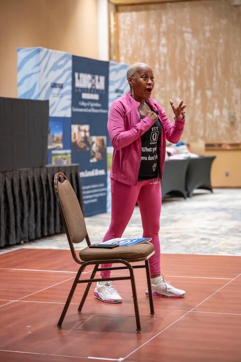 A woman in a pink tracksuit holds a microphone while standing next to a chair.