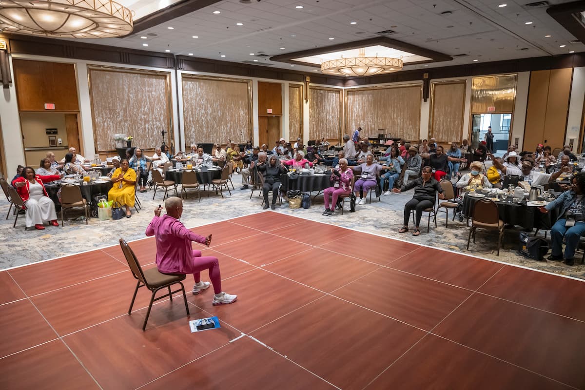 A woman in a pink exercise outfit leads a demontration in a conference room.