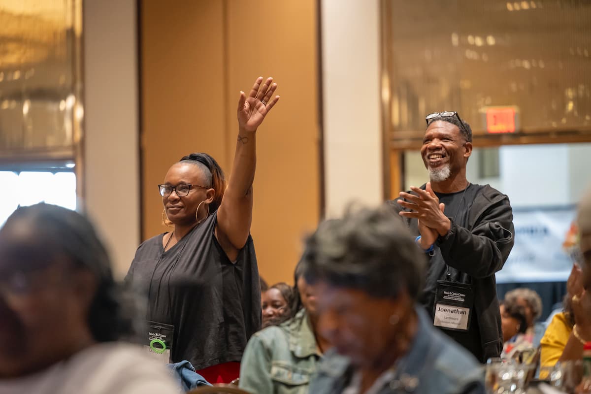 A woman raises her hand while standing in a conference room full of people sitting at tables.