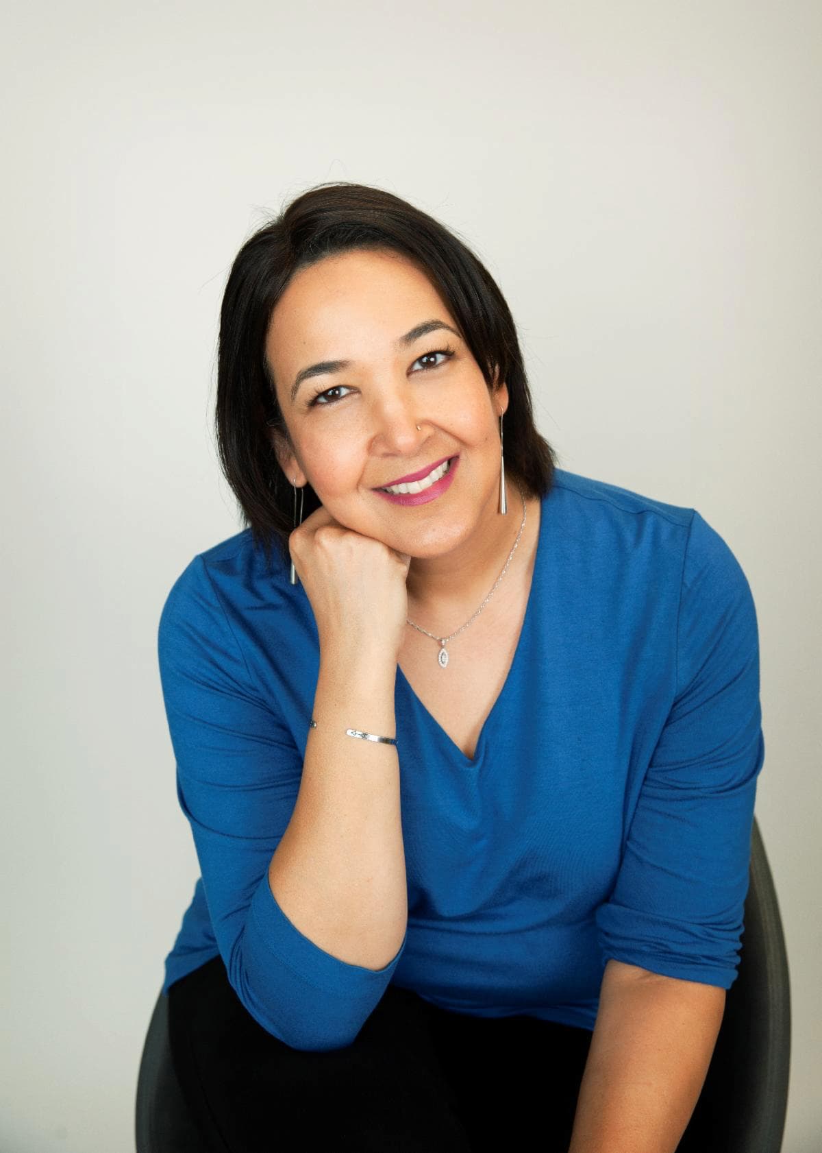 A woman with medium-length dark hair, wearing a blue top, sits smiling with her head resting gently on her hand. She has a nose stud, wears drop earrings, a delicate necklace, and a silver bracelet. The background is a plain light color.