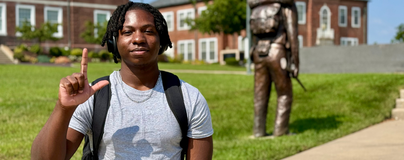 A Lincoln University student stands on campus making an “L” hand sign, with a historic statue and academic building in the background.