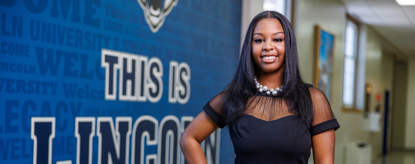 A Lincoln University student stands on campus making an “L” hand sign, with a historic statue and academic building in the background.