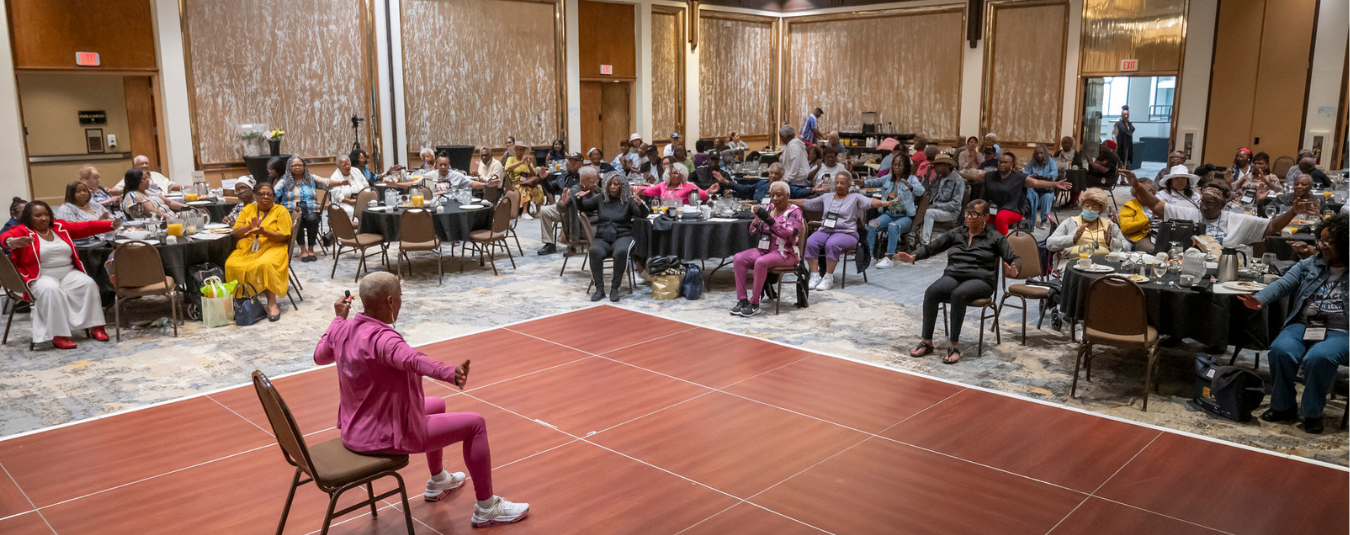 A woman in a pink tracksuit suits in a chair leading exercises at the minority institute of health and aging conference 2025.
