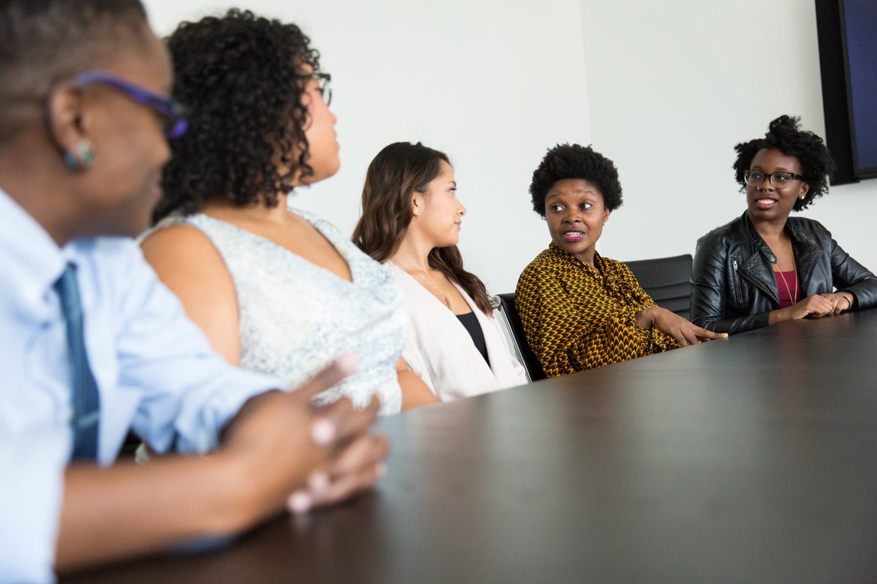  Five people seated at a conference table in discussion, with a Black woman in a yellow patterned blouse gesturing as she speaks to the group.