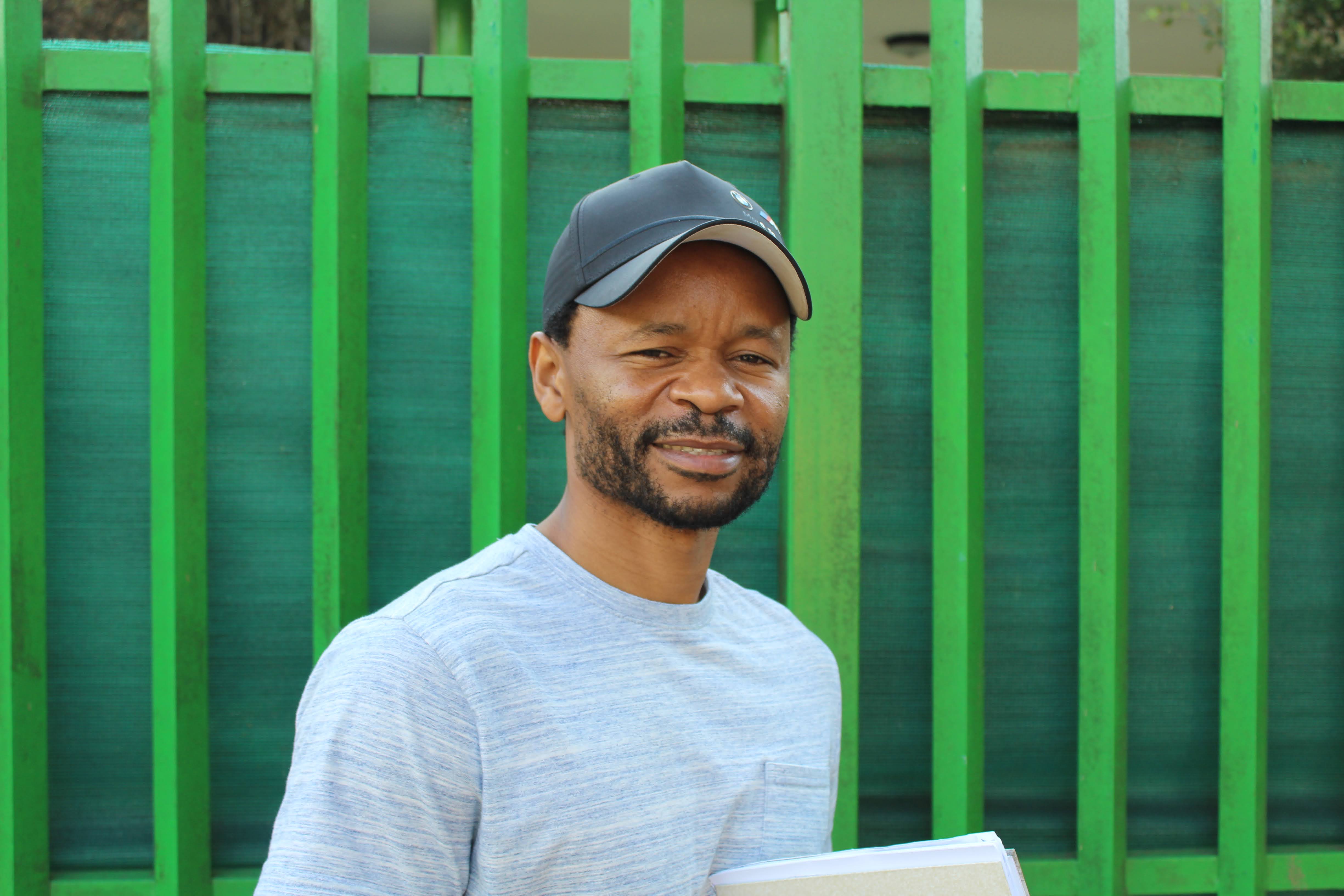Man in a light gray shirt and black cap smiling in front of a green fence, holding a folder or notebook.
