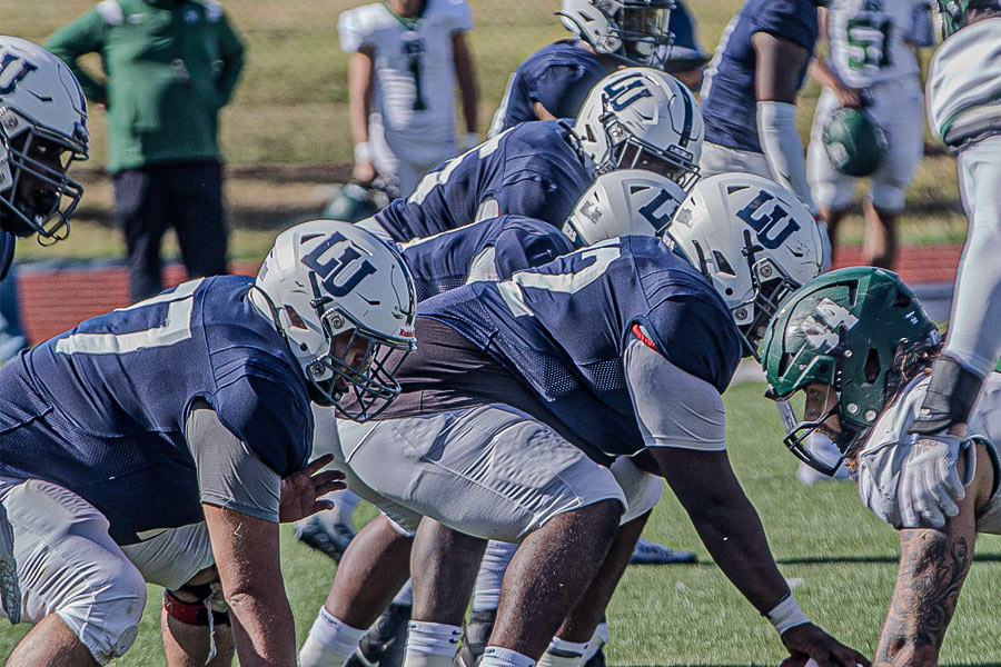 Lincoln University football players in blue and white uniforms line up on the field against an opposing team before the snap.