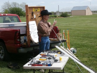 Person demonstrating electric fence tools beside a pickup truck, with fence posts, wires, and equipment laid out on a table.