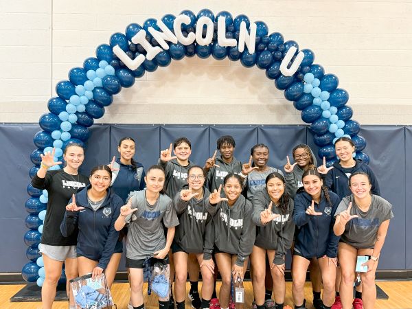 Group of Lincoln University students posing under a blue balloon arch reading “LINCOLN U” in a gym.