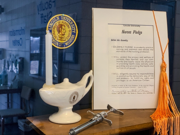 Display of nursing pinning items: candle, lamp, syringe, tassel, and framed Nurses Pledge from Lincoln University.