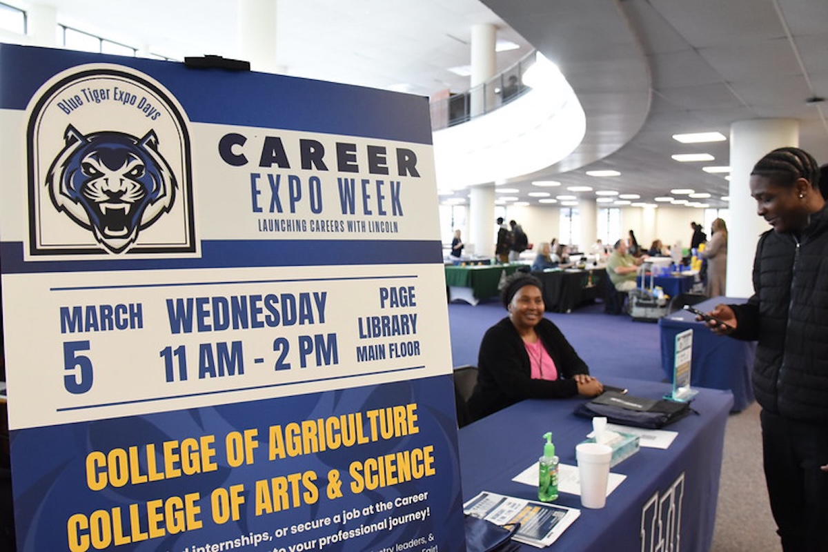 Career Expo Week event with a sign and booth promoting the College of Agriculture and Arts & Science. A person stands near the table where another person is seated, in a library setting.