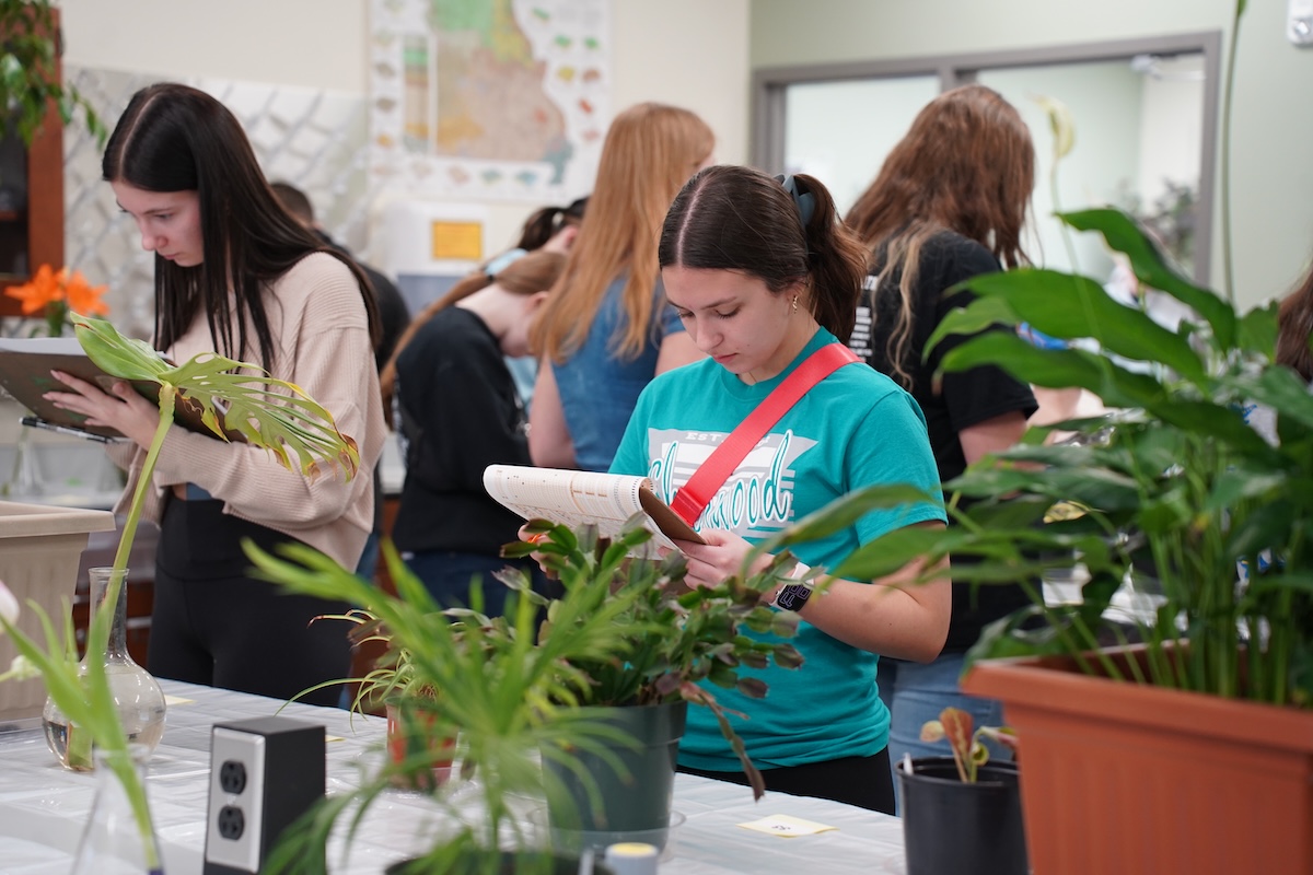 Several teenage girls with clipboards look at plants on a table.