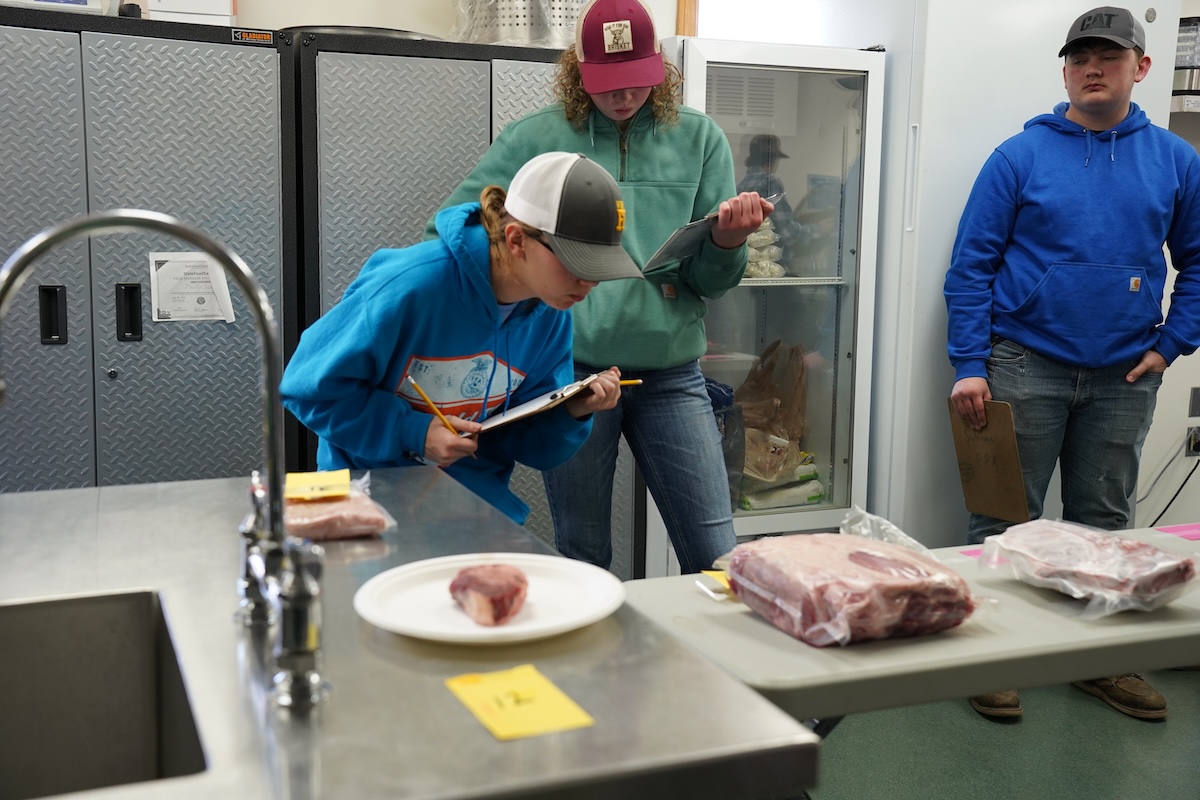 Three teenage students look at various meats in a kitchen setting.