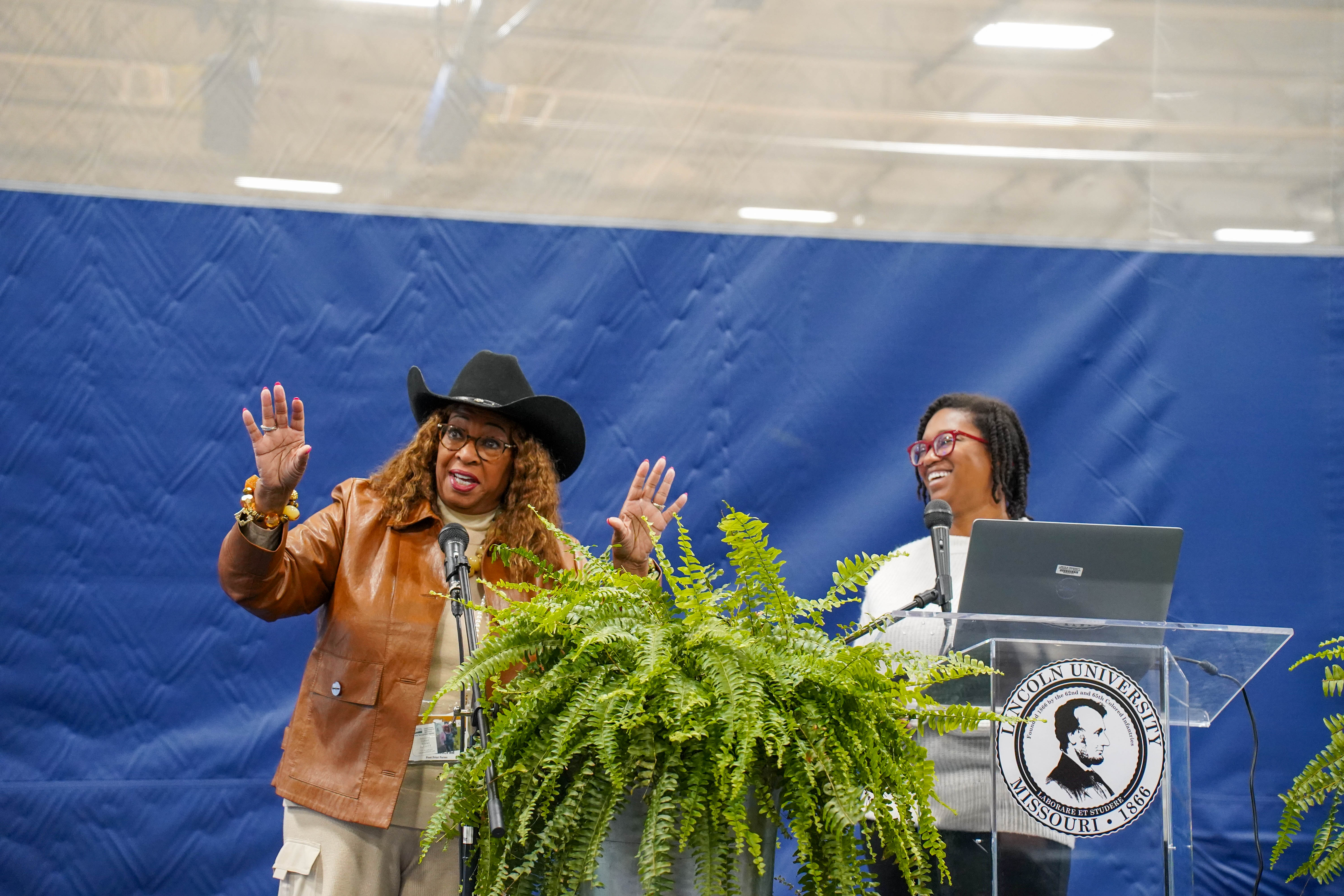 Two women stand speaking at a glass lectern with ferns on either side.