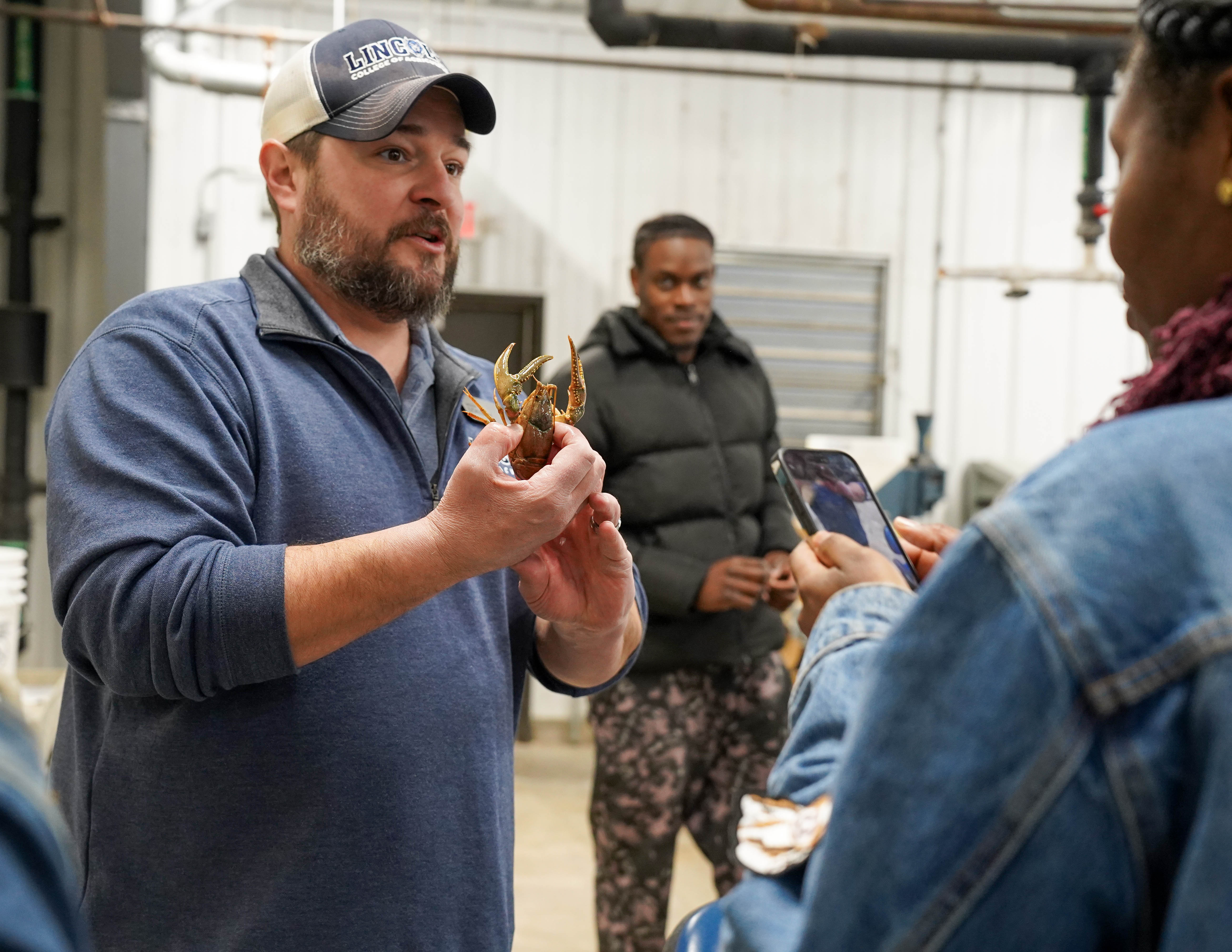 Two people stand watching as a man shows them a crawfish. 