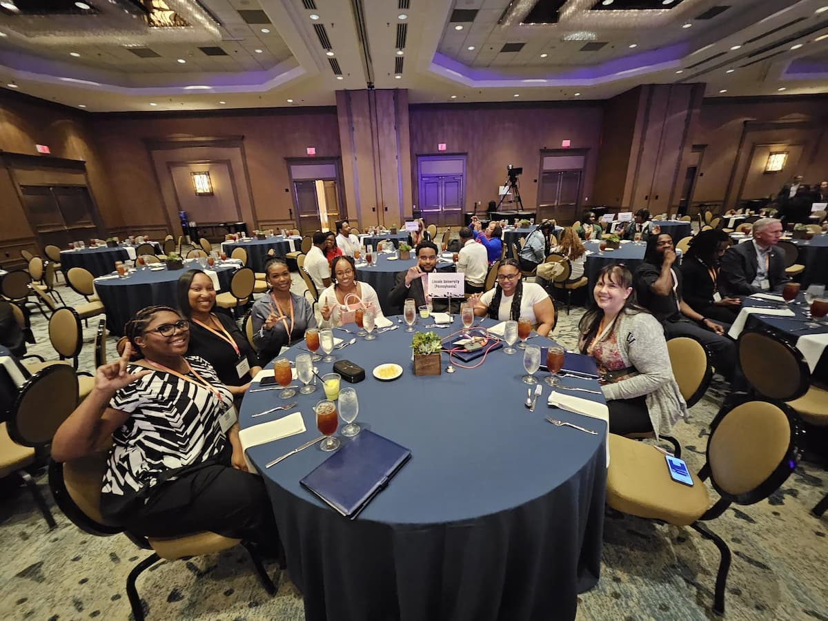 Lincoln University Strada Scholars and program representatives seated at a round banquet table during a conference luncheon. The table is set with drinks and name placards, and several attendees are smiling and making the 'L' hand sign for Lincoln.
