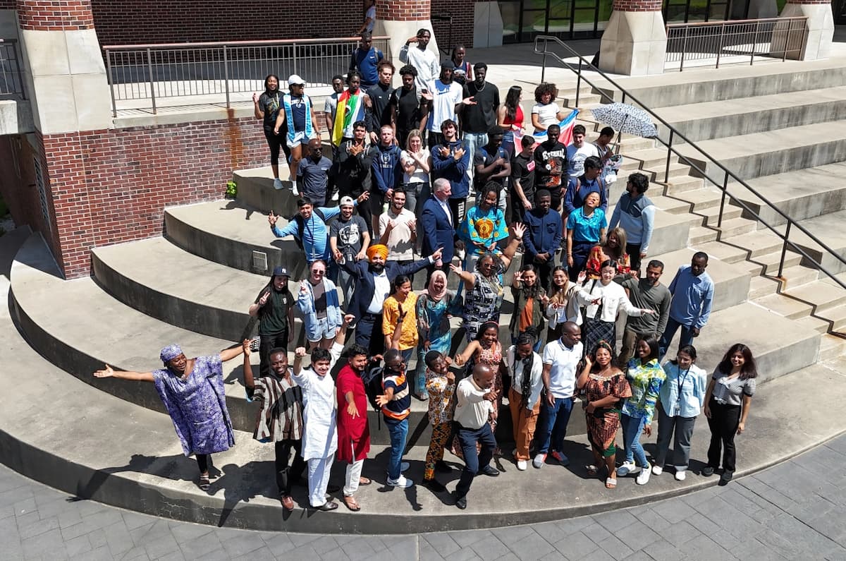 A vibrant group of international students arranged on circular concrete steps outside a brick building, some waving and smiling, others holding flags, showcasing cultural pride and unity on a sunny day.