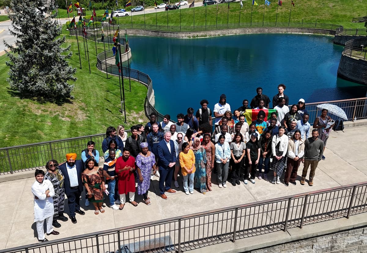 A group of international students and faculty members standing on a terrace in front of a pond with international flags, posing together under bright sunlight with a mix of colorful traditional and modern clothing.