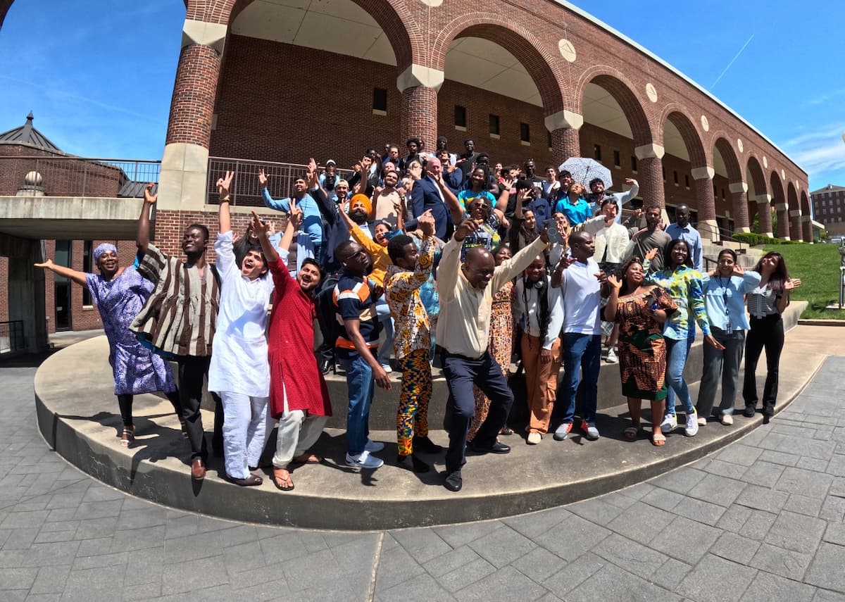 A vibrant group of international students arranged on circular concrete steps outside a brick building, some waving and smiling, others holding flags, showcasing cultural pride and unity on a sunny day.