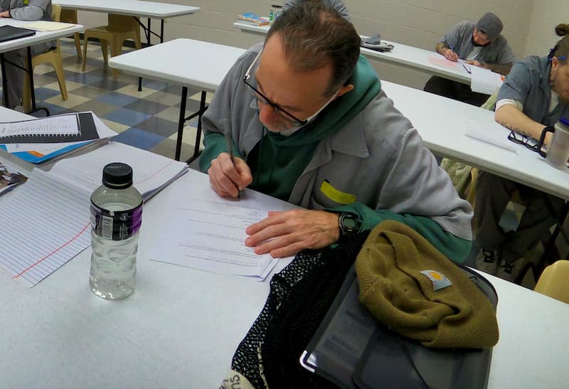 "A man wearing glasses and prison uniform is focused on writing at a desk in a classroom setting. Other individuals in similar attire are also seated at tables, working on papers. The room has white tables, plastic chairs, and a tiled floor.