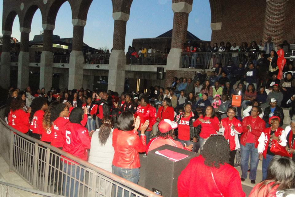 A large group of women, many wearing coordinated red and white jackets with Greek letters, gather in a semicircle at an outdoor amphitheater. The crowd appears to be part of a Delta Sigma Theta sorority event, as indicated by the Greek letters ΔΣΘ on their clothing. Spectators fill the surrounding steps and upper level, watching and cheering. The atmosphere is lively and communal, with a mix of applause and engagement.