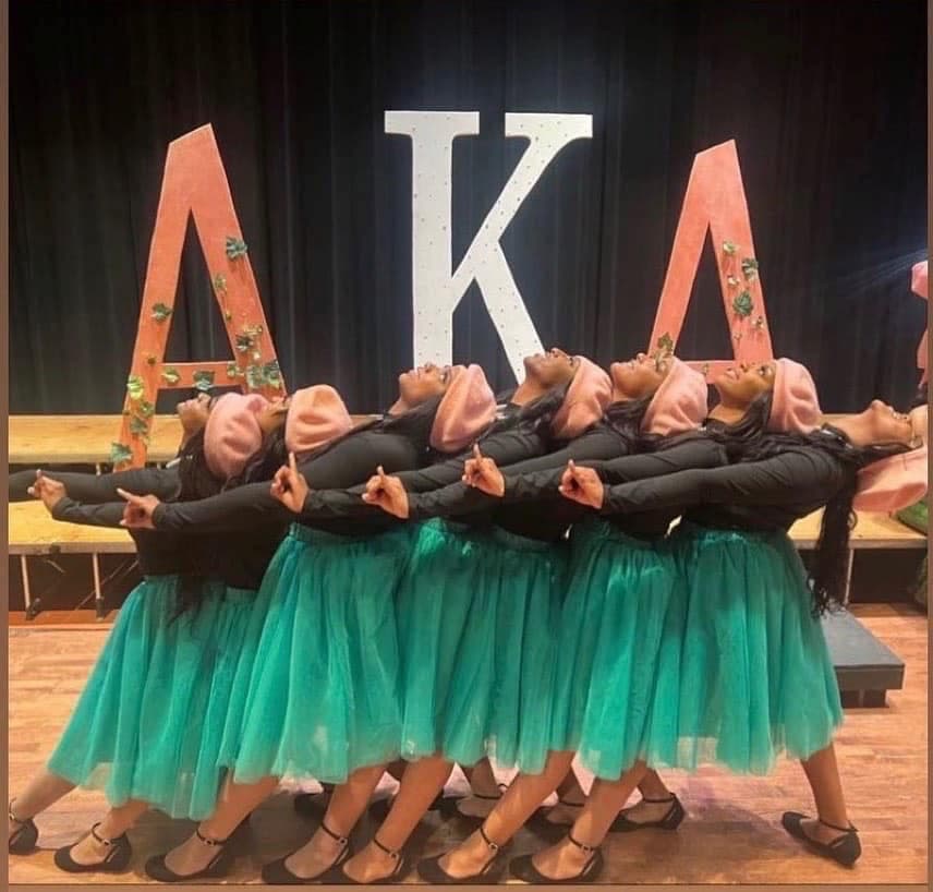 Lincoln University of Missouri AKA sorority members pose in a line for a photo. They are dressed in green skirts, black shits and pink berets. 