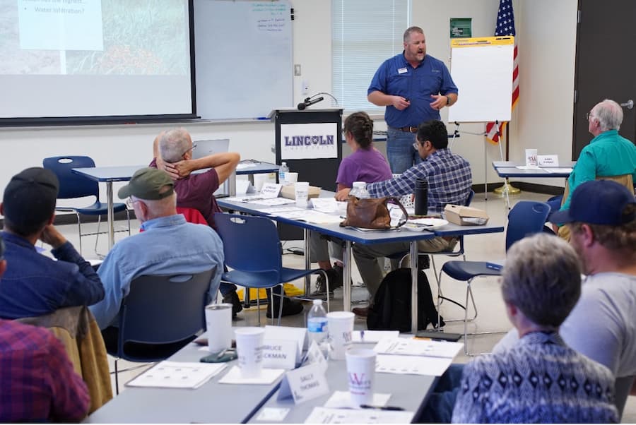 A man stands in front of a classroom full of people. To his left, a projector shows a slideshow presentation. 