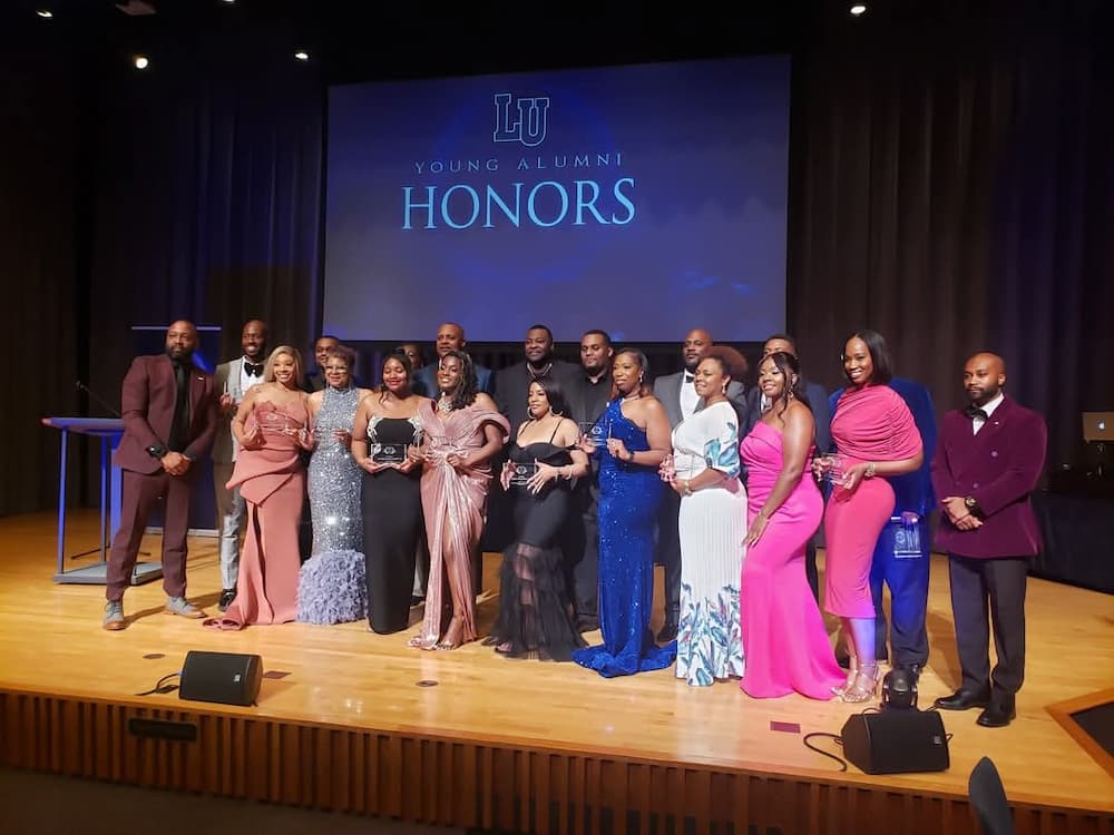 A group of elegantly dressed award recipients and organizers stand on stage at the Lincoln University Young Alumni Honors Awards. A screen in the background displays the event logo. The group smiles proudly while holding glass awards.