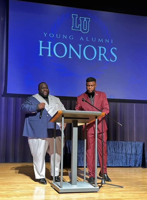 Two men stand on the stage at Unity Village behind a podium during the 2025 Young Alumni Awards Honors. 